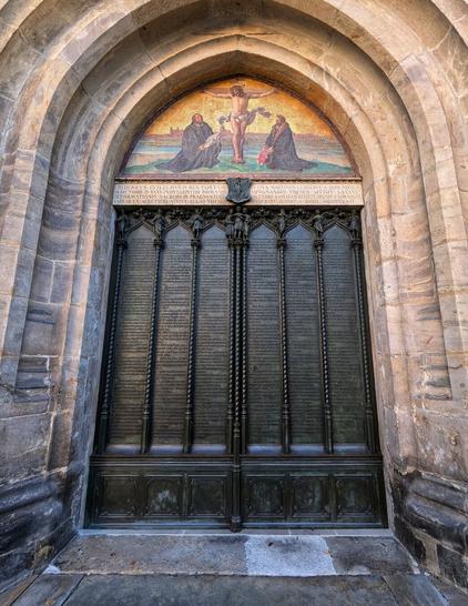 The image shows the "Theses Doors", the entrance to the Church of All Saints in Wittenberg, Germany. The entrance is framed by a large, arched stone doorway. Above the door, there's a mosaic depicting the crucifixion of Jesus, flanked by two figures. The door itself is made of dark bronze and features intricate carvings and inscriptions, commemorating Martin Luther's 95 theses. The stone surrounding the doorway shows signs of weathering, adding to the historical feel of the scene.