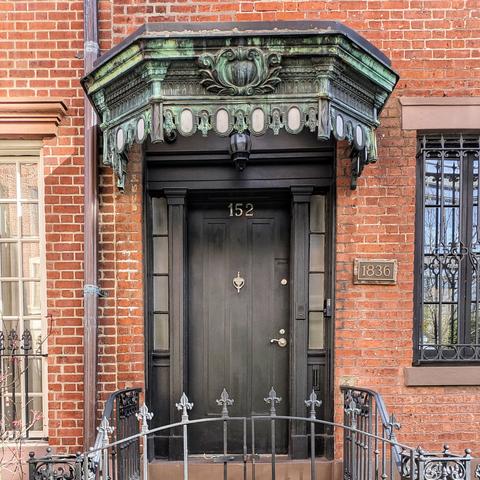 Ornate entrance of a historic brick building with a dark door numbered "152" under a green metal canopy, a bronze plaque reading "1836", and a black wrought-iron railing.