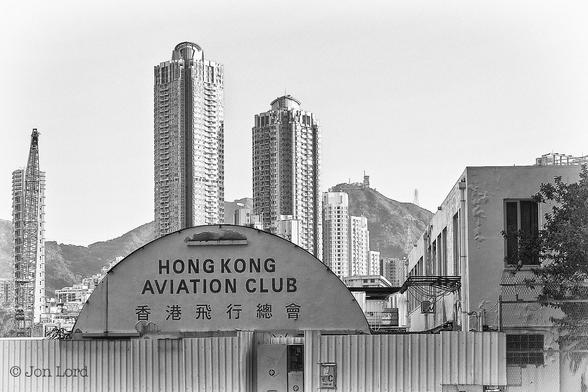 This Is A Black And White Cityscape Photo In Landscape Format. Running Along The Base Of The Image And Rising Up Only About Ten Per Cent Is A White Corrugated Fence Finishing In The Lower Right Corner Where There Is A Short Length Of Tall Wire Fence With A Long (Stretching Away From The Camera), White Two Story Building. Below And To The Left Of Centre And Behind The White Corrugated Fence Is A Weathered White Semicircular Building, No More Than Three Metres Tall And Perfectly Aligned With The Camera So The Sides Are Not Visible. On This Visible End Of The Building Are The Words 'Hong Kong Aviation Club' In Large Bold Black Letters And 香港飛行總會 Below. In The Middle Ground Behind The First Two Buildings Are A Number, About Ten, Of Very High-Rise Apartment Buildings, All White. In The Background Stretching From Left To Right Is A Ridge Of Undulating Mountainous Terrain With A Bland White Sky Above. 
The Location Is Kowloon, At The Site Of The Former Kai Tak Airport In Hong Kong (2016).