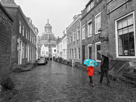 A monochrome street scene shows two people with umbrellas walking away from the viewer. The child on the left is selectively coloured, wearing a bright red coat and holding a blue umbrella with colourful designs. The adult on the right and the rest of the scene are in black and white.