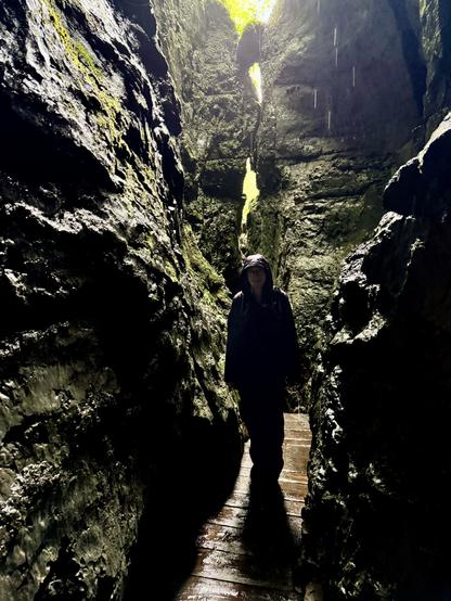 Person in shadow within narrow part of gorge, standing on wooden ledge above stream bed; water droplets indicate rainfall