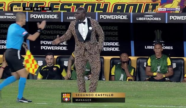 A man dressed in a leopard print suit stands on the side of a football pitch at the beginning of a game ready to shake hands with an official