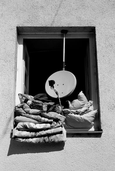 The black and white image shows an open window with a satellite dish mounted above it and several thick blankets and pillows stacked on the windowsill, spilling slightly outward.