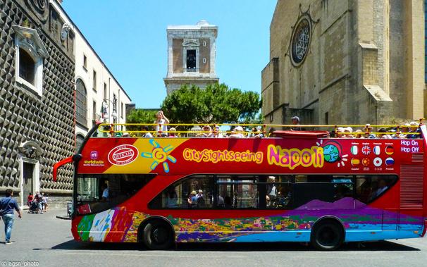 Seitenansicht eines vollbesetzten Sightseeing-Busses auf der Piazza del Gesù Nuovo in Neapel. Touristen mit Sonnenhüten sitzen auf dem offenen Oberdeck und fotografieren den Platz. /
Side view of a sightseeing bus on the Piazza del Gesù Nuovo in Naples. Tourists wearing sun hats sit on the open upper deck and photograph the square.