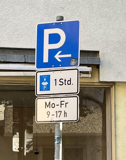 Schild mit Hinweis, dass zwischen 9:00 und 17:00 Uhr eine Stunde geparkt werden darf mit Parkscheibe. Dahinter ein Schaufenster, in dem sich die gegenüber liegende Häuserzeile spiegelt.