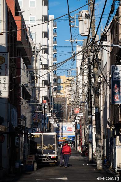 This photograph captures a vivid urban moment in the backstreets of Asakusa, Tokyo.

It reveals a city crisscrossed with overhead wires, casting a dense net above narrow alleyways and silhouetted figures.

Framed by apartment blocks and flanked by signage, utility poles, and delivery trucks, the scene captures the authentic pulse of Tokyo life on a crisp morning. Sunlight slices through the narrow passage, highlighting textures and contrasts that tell stories of movement, routine, and quiet resilience.

Ideal for interiors that embrace raw urban aesthetics, documentary-style photography, or love the organized chaos of Japanese cityscapes.

Ideal for collectors of Tokyo street photography, lovers of Japanese architecture, and fans of urban geometry and everyday life scenes.