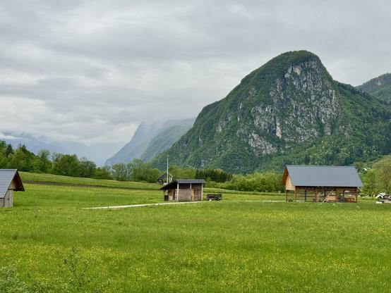 View from motorhome parking area (Stellplatz) just outside village of Srednja Vas v Bohinju. Meadow foreground with several small farm-related structures; mountains in background.