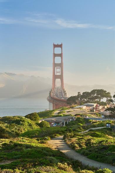 A view from the south looking along the span of the Golden Gate Bridge. The foreground is a brilliant green. The Bridge towers are red. The far tower is visible through the close tower. There is a wisp of fog coming from west slightly obscuring the Marin Headlands. The whole scene is bathed in early morning sunlight.