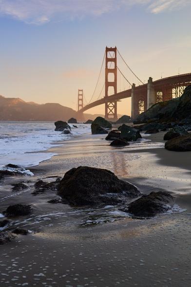 A sunrise view of the Golden Gate Bridge from a beach just to the southwest. The Gate is lit with the first sunlight. The beach is in shadow and the surf is reflecting the ambient light. The foreground includes many boulders in the surf.