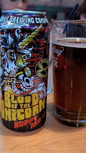 On the left: can of Pipeworks Brewing Blood of The Unicorn Hoppy Red. On the right: a glass full of dark amber beer.