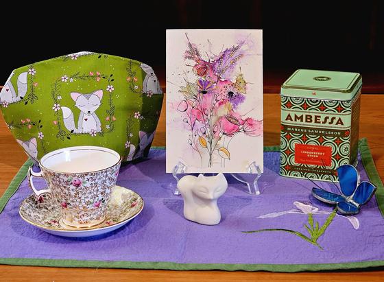 A springtime tea tableau in light purple and green with a fox patterned cozy, a bisque porcelain Art Deco style fox, a blue stained glass butterfly, an abstract watercolor painting, vintage china, and Lingonberry green tea.