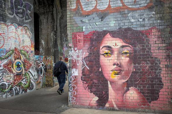 The main focus of the picture is a street art painting of a woman's face on a brick wall. Another street art piece is visible on an opposite wall, a triffid like creature. A path goes between the walls. A figure is walking between, with their back visible. The path between the walls shows that the walls are actually part of very tall, brick arches.