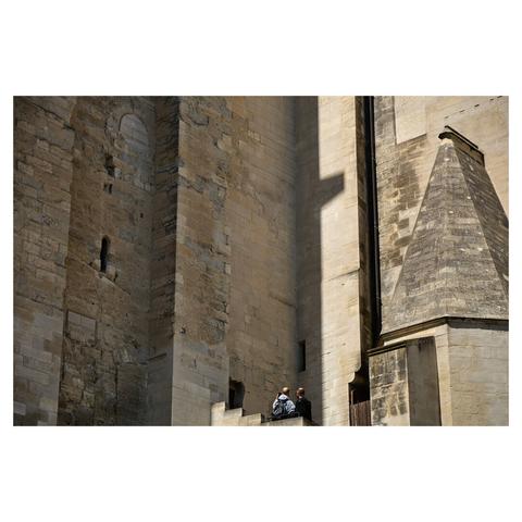 Taken at the inner court of Palais de Papes in Avignon, the image shows the massive walls of the palace rising up, filling the entire frame. At the center bottom, two tourists are standing on a staircase with their backs to the camera, taking a selfie.
