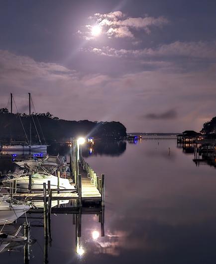 Night view over a waterway's merge into a larger river, with lighted shorelines lined with sailboats and boat docks, with a glowing moon overhead casting a reflection upon the water below.
