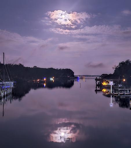 Night view over a waterway's merge into a larger river, with lighted shorelines lined with sailboats and boat docks, with a glowing moon overhead casting a reflection upon the water below.