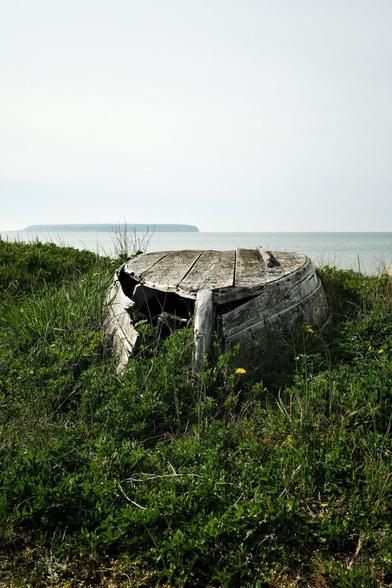 En gammal upp och nedvänd träbåt bara för har spruckit, ligger uppdragen i gräset. I bakgrunden syns havet och en ö i soldiset.