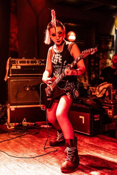A guitarist with a punk-inspired hairstyle performs on stage, wearing a sleeveless black shirt and denim shorts. The setting is dimly lit, with visible amplifiers and musical equipment in the background. The atmosphere suggests an energetic live music performance.