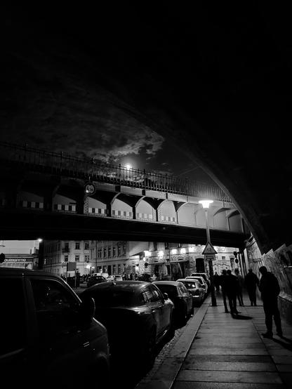 Das Bild zeigt eine nächtliche Szene in einer Stadt, aufgenommen von unter einer Brücke aus. Die Brücke ist aus Metall und hat mehrere Bögen, die durch die Dunkelheit erahnt werden. Auf der Brücke sind einige Personen zu sehen, die sich auf dem Gehweg unter ihr bewegen. Die Straßenlaternen werfen ein warmes Licht auf die Szene, das die Umgebung in ein schwarz-weißes Bild taucht. Parkierte Autos säumen die Straße, und im Hintergrund sind Gebäude mit Fenstern und Details zu erkennen. Der Himmel ist dunkel, mit einer sichtbaren Mondlichtung, die durch die Wolken hindurchscheint. Die gesamte Atmosphäre wirkt ruhig und stimmungsvoll, mit einem Hauch von urbaner Nachtlebensatmosphäre.