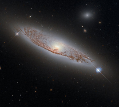 Galaxy NGC 5037 seen edge-on with a bright core and sweeping reddish dust lanes, captured by the Hubble Space Telescope. A foreground star with diffraction spikes appears on the right, and distant galaxies dot the background.