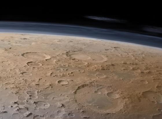 A digitally colourised image of Mars Promethei Terra region, captured by ESA’s Mars Express from orbit, shows a vast, cratered landscape near the massive Hellas Planitia basin. Dominating the scene are three major impact craters: Secchi Crater with a wide, sharply defined rim, Wallace Crater partially overlapping nearby craters and the more eroded Tikhov Crater with softened edges. The surface is textured with countless smaller craters, in tones of ochre, red, and dusty brown, evoking a parched desert. Above the horizon, a thin bluish layer of Mars atmosphere fades into deep black space. Wispy white clouds stretch across the upper layer of the atmospheric limb, offering a glimpse of Martian weather. The planet’s curvature is visible.