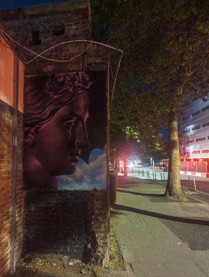 On a street at night, a wall has a large street art painting of a greek goddess (or statue of such) in red with a black and blue background.