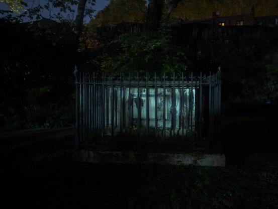 At night, a tomb is surrounded by spiked railings. There's a tall hedge behind.