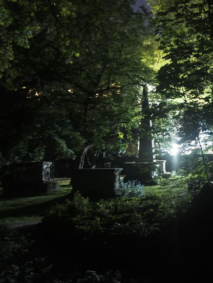 View through a graveyard at night. There's several tombs, a tall obelisk, and between and behind them trees and bushes. Behind a wall is a bright light.