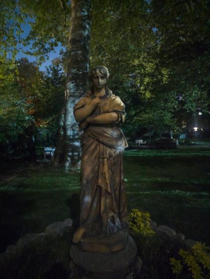 View of a statue of a woman wearing a robe, in a park at night. The statue is missing one hand.