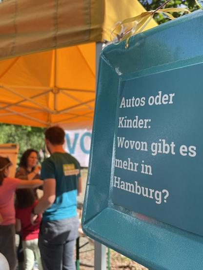 Im Vordergrund ist ein Schild von einem Ratespiel mit der Frage: "Autos oder Kinder: Wovon gibt es mehr in Hamburg?". Im Hintergrund sind Menschen zu sehen, die sich unterhalten.