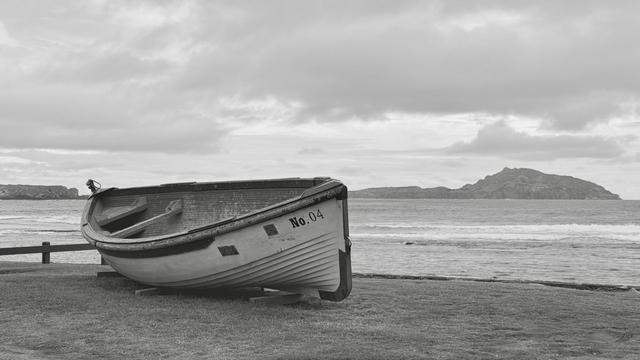 Black and white image of a wooden boat sitting on an ocean shore. Behind it are calm ocean waters and in the distance a rocky island is visible.