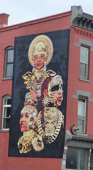 A Mural on Main Street in Buffalo, NY. The photographer is unaware of the creator or symbology. Three golden and fabric ordained portraits of Afro-futuristic design. The lowest head is facing left with a smile and eyes looking at the camera. Her headdress has orange, light-blue, and white fabric. Her ears to chest are adorned in gold. There is a white line of face paint from her lower lip down her chin. A man is looking right located above her shoulder. His sunglasses are framed by gold adornment, also extending in vibrant patterning down his chest. Behind his head and above him, facing above the photographer forward, has a circular golden headdress with circular patterns to its center and across her forehead. Her earrings have three baubles. Her shoulders have black and gold triangles, ten in total. Her chest has a purple, blue, white, golden breastplate or fabric dress. They are framed by a dark black square background, with darker shaded leaves of different plant varieties.
