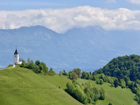 View of church atop meadow with alps in background, seen from Jamnik village