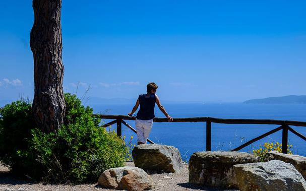 Eine Frau steht auf einer Aussichtsplattform, lehnt an der Holzbarriere und blickt auf das blaue Meer. / 
A woman stands on a viewing platform, leaning on the wooden barrier and looking out at the blue sea.