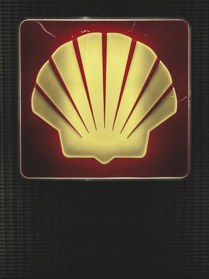 Illuminated Shell logo sign featuring a bold yellow scallop shell with red lines on a deep red background, mounted against a dark grid-textured wall.