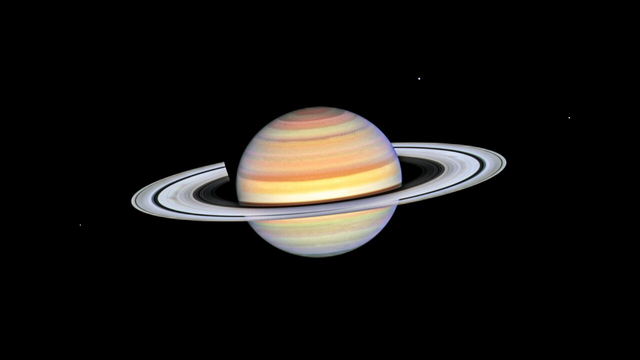 From source in toot: Planet Saturn with bright white rings, multi-colored main sphere, and moons Mimas, Dione, and Enceladus. Spoke features on the left and right sides of the rings appear like faint grey smudges against the rings’ bright backdrop, about midway from the planet to the rings’ outer edge. Above the rings plane, the planet’s bands are shades of red, orange and yellow, with bright white nearer the equator