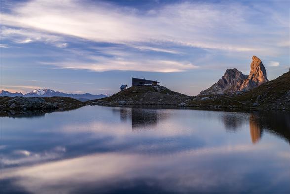 Photo d'un paysage de montagne au lever du soleil.
Au premier plan on voit l'eau d'un lac troublée par une légère brise. L'horizon est fermé par la rive opposée, dominée par un refuge de montagne au milieu et un pic montagneux à droite déjà éclairé par le soleil. 
Tout au fond à gauche, on distingue des montagnes au loin, l'une d'elles est enneigée et aussi éclairée par le soleil.
-
Photo of a mountain landscape at sunrise.
In the foreground, the water of a lake is disturbed by a gentle breeze. The horizon is closed by the opposite shore, dominated by a mountain hut in the middle and a mountain peak on the right, already illuminated by the sun. 
In the far left, we can make out mountains in the distance, one of them snow-capped and also lit by the sun.