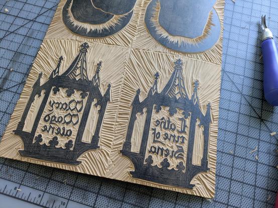 A partially carved linoleum block on a cutting mat. The design shows two "church-shaped" reliquary-like frames around text in a gothic script (based on Bastarda, for anyone interested). The text is backwards and reads "Mary Magdalene" on one side and "Catherine of Siena" on the other.
