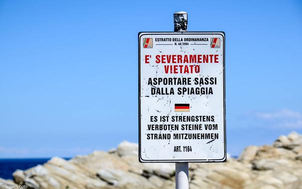 Nahaufnahme eines zweisprachigen Verbotsschildes am Strand der Insel Elba. Text auf Italienisch und Deutsch darunter: Es ist strengstens verboten, Steine ​​vom Strand mitzunehmen. /
Close-up of a bilingual prohibition sign on the beach on the island of Elba. Text in Italian and German below: It is strictly forbidden to take stones from the beach.