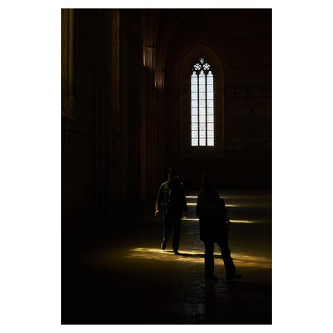 Taken in a large hall at Palais des Papes in Avignon. The image is mostly dark, with a few patches of light coming through the stained windows. A visitor is stepping into the light, another person is standing next to them in the dark with only their silhouette visible.