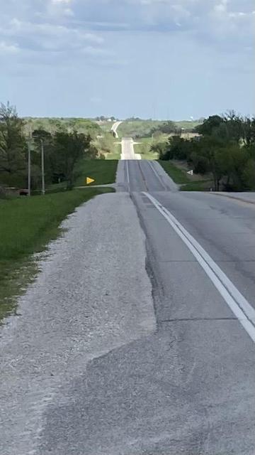 A two-lane rural highway climbs and descends rolling green hills and drifts from side to side.  The far horizon is about 8 miles (13 km) in the distance.