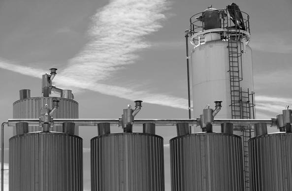 The image shows a black and white photo of industrial tanks or silos. There are several large cylindrical containers with pipes and vents on top. In the background, there is a taller vertical structure with a ladder attached to its side. The sky is visible with some cloud patterns.