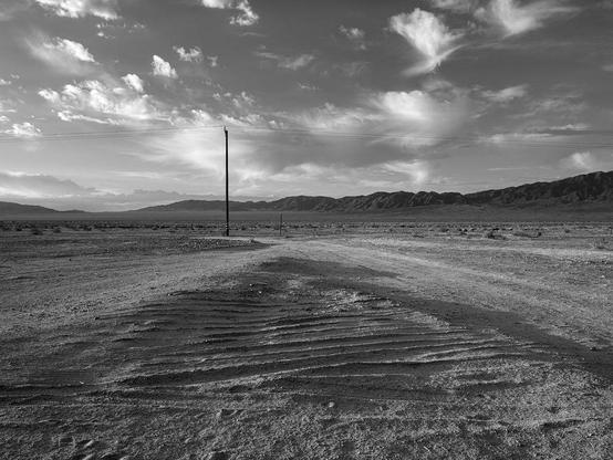 Desert landscape with wheel ruts in the foreground, power line in the middle, and mountains with partly cloudy sky in the background.