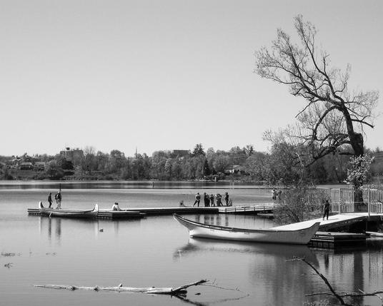 Large canoes sit next to wood docks on a lake, with people standing on the dock. 
The calm water of the lake reflects nearby trees with the distant shoreline seen in the background and a very large leafless tree seen on the right. This is a black and white image.