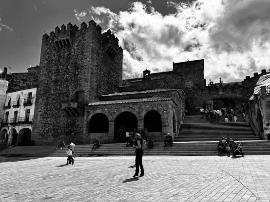Una fotografía en blanco y negro muestra una plaza adoquinada en una ciudad histórica, dominada por una imponente estructura de piedra con una torre alta y almenada.  Escaleras anchas suben a la estructura, flanqueadas por arcos. Varias personas se encuentran en la plaza, algunas sentadas en los escalones, otras caminando. El cielo está parcialmente nublado. La arquitectura es de estilo antiguo, con muros de piedra gruesa y detalles arquitectónicos visibles.  La escena transmite una sensación de historia y serenidad.