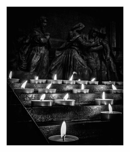 A black and white photo taken from inside a church. In the foreground there is the the stand that people put little tea light candles on which are used for prayers. There is one candle in the middle of the photo right at the bottom with four rows behind it with other candles scattered. In the background you can make out the highlights of four of the statues from Lombardi's Compianto. They are all wearing loose fabrics which give lots of movement and texture. Two figures are holding on to/back a third which seems to be in great distress. The fourth has their hands clasped together and appears to be praying.