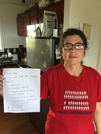 Beth Mazur wearing a red Millions MIssing shirit and holding up a piece of paper saying she has had myalgic encephalomyelitis/chronic fatigue syndrome for 9 years and it has prevented her from living anything like a full and rich life.