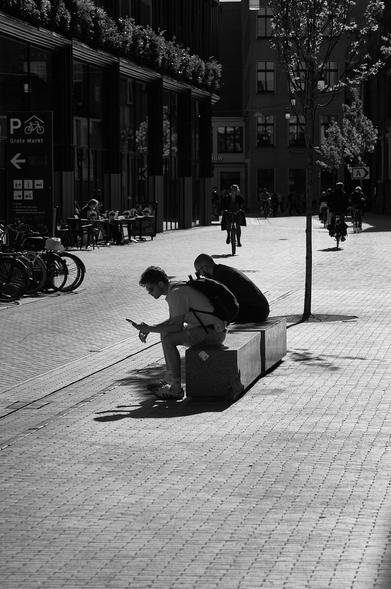 A black-and-white street scene showcases two seated individuals on a bench, one looking at a smartphone. In the background, people ride bicycles and dine at outdoor tables. Trees and modern buildings frame the scene, creating a lively urban atmosphere.