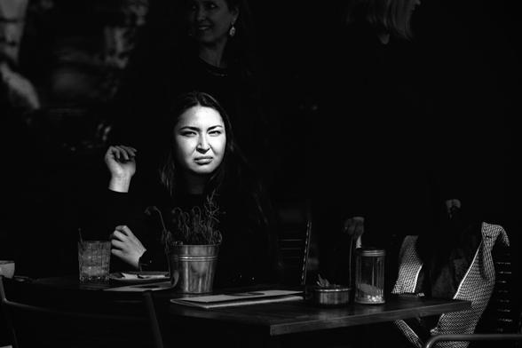 A medium shot captures a person with long, dark hair sitting at a table outdoors. The person is wearing a dark long-sleeved shirt and looking directly at the viewer with a not happy expression. Their left arm rests on the table with their hand near their face, while their right arm is also on the table.

On the wooden table in front of them, there is a small metal pot containing green plants, a clear glass with ice and a straw, a white plate with cutlery, and a small metal container. The table is partially illuminated by sunlight, creating strong contrasts of light and shadow.

Behind the person, two other individuals are partially visible and out of focus. One person is wearing a red jacket and standing, while the other is wearing a dark garment and is also standing. The background is dark and indistinct, suggesting a dimly lit outdoor setting. The focus is sharply on the person sitting at the table, drawing the viewer's attention to them.