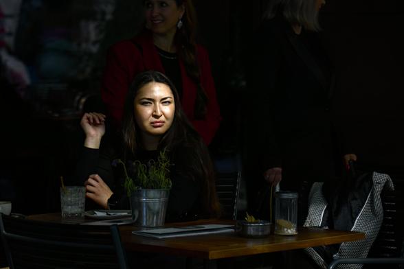 A medium shot captures a person with long, dark hair sitting at a table outdoors. The person is wearing a dark long-sleeved shirt and looking directly at the viewer with a not so happy expression. Their left arm rests on the table with their hand near their face, while their right arm is also on the table.

On the wooden table in front of them, there is a small metal pot containing green plants, a clear glass with ice and a straw, a white plate with cutlery, and a small metal container. The table is partially illuminated by sunlight, creating strong contrasts of light and shadow.

Behind the person, two other individuals are partially visible and out of focus. One person is wearing a red jacket and standing, while the other is wearing a dark garment and is also standing. The background is dark and indistinct, suggesting a dimly lit outdoor setting. The focus is sharply on the person sitting at the table, drawing the viewer's attention to them.