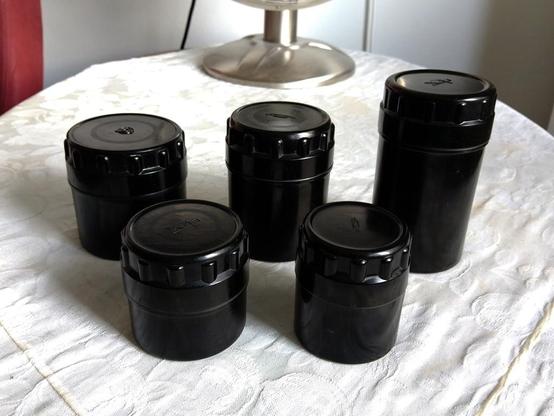 Five black, round bakelite containers used for storing Soviet-made lenses and other peripherials, on a table.